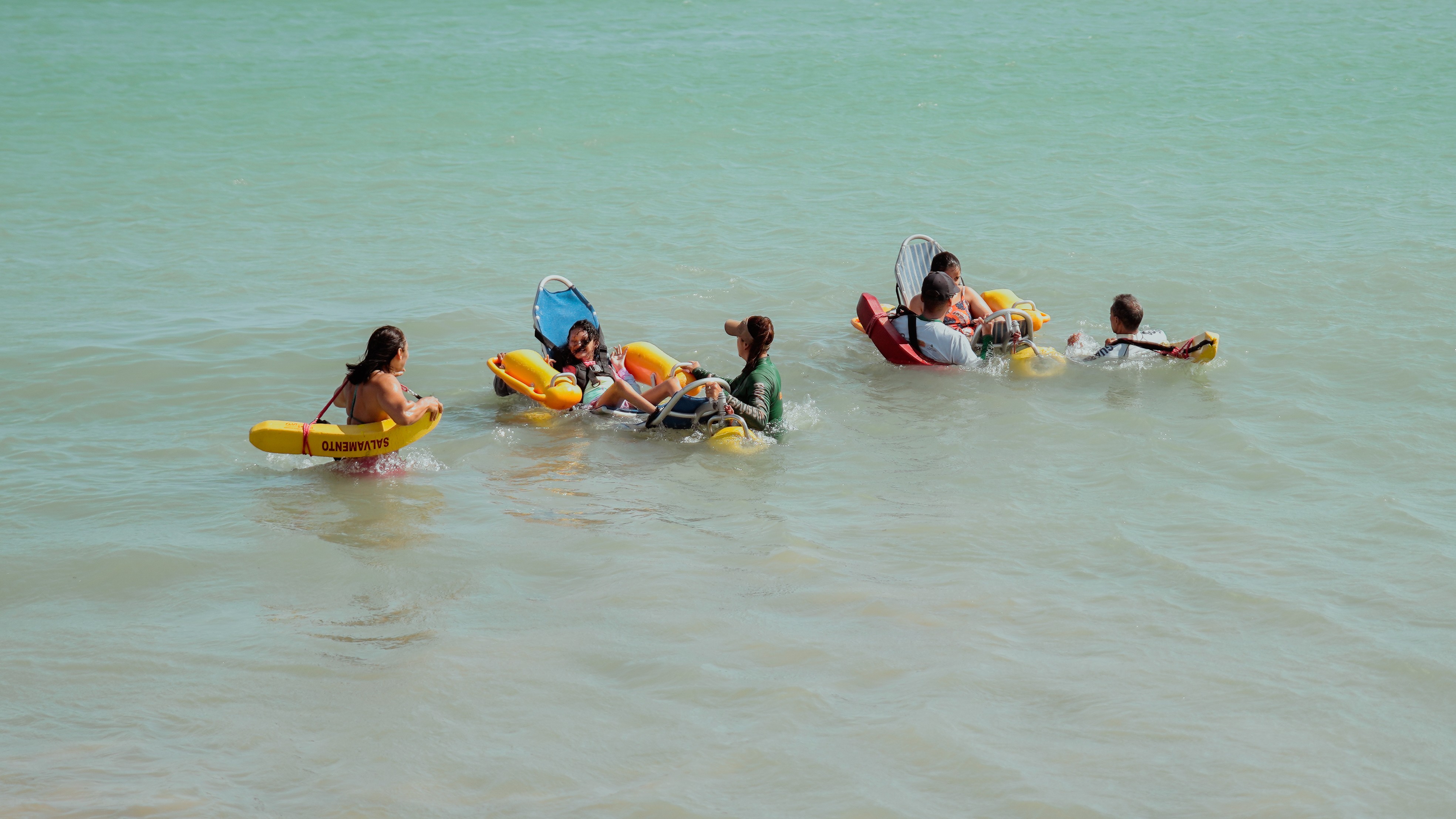 Duas crianças em cadeiras anfíbias, auxiliadas por salva-vidas, tomam banho de mar